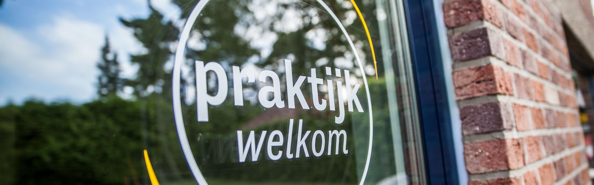 De raamsticker van de praktijk met de tekst praktijk welkom in een okeren cirkel, op het raam naast de bakstenen gevel.